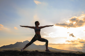 Silhouette of young woman practicing yoga or pilates at sunset or sunrise in beautiful mountain location, doing lunge exercise, standing in Warrior.