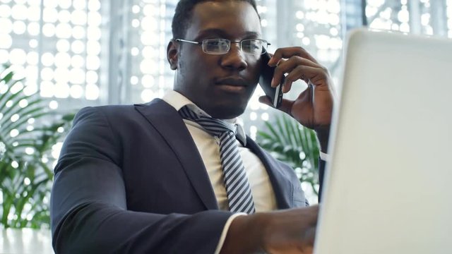 Close-up shot of intelligent African American businessman in eyeglasses typing on laptop and talking on mobile phone in coworking space