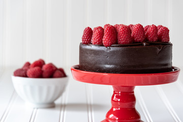 Chocolate cake with red raspberries on top, on a red cake platter, and red raspberries in a white...