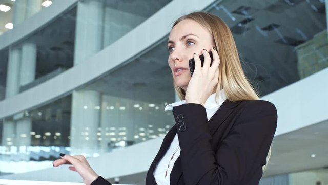 Attractive caucasian businesswoman in formal suit standing in large modern office building and talking on mobile phone