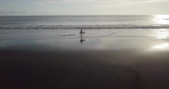 Fit surfer woman in sexy bikini and short shorts with white surfing board standing on a black beach at sunrise. Modern lifestyle. Sport camp and extreme swim on summer vacation. Aerial drone footage.