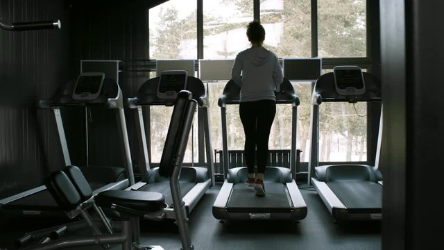 Rear view of young sportswoman running on treadmill in empty gym and looking out of windows