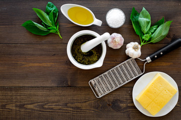 Ingredients for pesto sauce. Cheese, garlic, green basil, olive oil, salt near grater and mortar on dark wooden background top view copy space