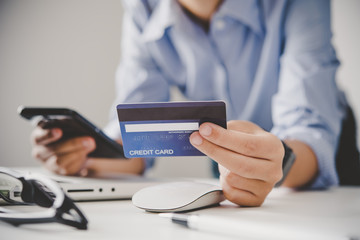 Fototapeta premium Hand holding a credit card in their hands and find information about a product using their mobile device to make purchases online and conduct financial transactions.