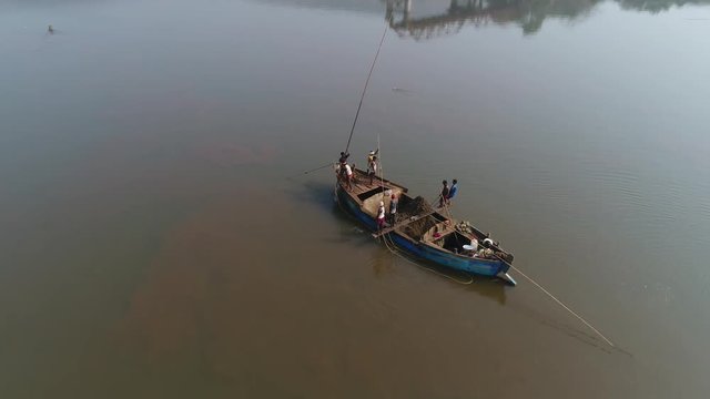 Workers with long poles standing on a wooden boat mined from the bottom of the river sand and clay for construction. Hard manual labor of Indian men. Aerial vew.