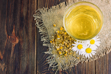 Glass of herbal tea with chamomile flowers on dark wooden background.