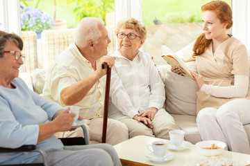 Tea time for seniors sitting on a couch in a common room of a luxury retirement home. Caretaker reading a book to elderly.