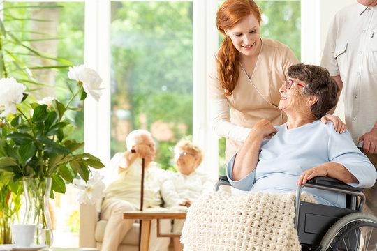 Tender caregiver saying goodbye to an elderly pensioner in a wheelchair in a day care facility.  A companion pushing the wheelchair. Other elderly people in the blurred background.