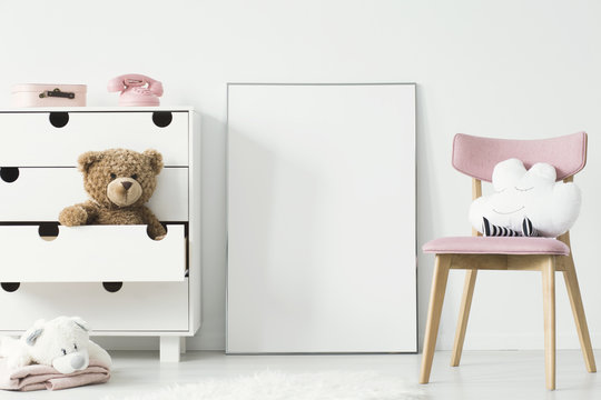 Empty poster with place for your picture standing on the floor in bright kid room interior with white cupboard and pink chair