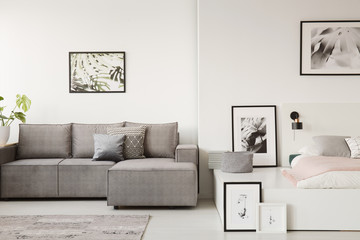 Real photo of a grey sofa with cushions standing next to a white platform bed in monochromatic...
