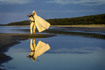 Portrait of assassin in white costume with the sword at the sea. He is posing near water with...