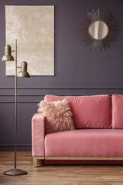 Furry pillow on a powder pink sofa, elegant golden sunburst mirror and marble painting on a dark gray wall in a living room interior