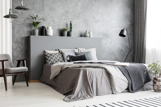 Stylish standing lamp next to a bed in a monochromatic grey bedroom interior with a footstool, an armchair and decorative plants in pots against the wall. Real photo.