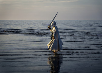 Portrait of assassin in white costume with the sword at the sea. He is posing near water during...