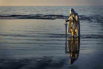 Portrait of assassin in white costume with the sword at the sea. He is posing near water during...