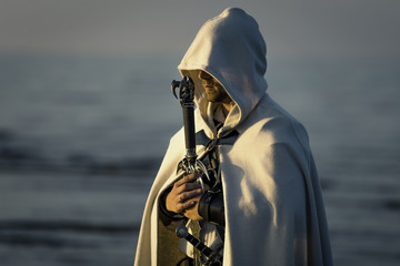 Portrait of assassin in white costume with the sword at the sea. He is posing near water during sunset, soft light. - 212038847