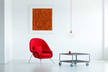 Real photo of a red armchair standing next to a table on wheels in a living room interior with...