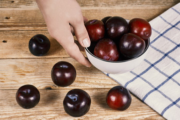 female hand holds a bowl with red plums next to a doily and a few plums in front, on a wooden background