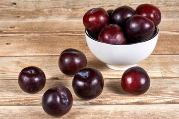 Red plums in a white bowl and a few plums in front, on a wooden background
