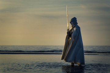 Portrait of assassin in white costume with the sword at the sea. He is posing near water during sunset, soft light. - 212039894