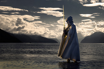 Portrait of assassin in white costume with the sword at the sea. He is posing near water during sunset, soft light. - 212040041