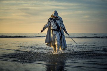 Portrait of assassin in white costume with the sword at the sea. He is posing near water during sunset, soft light. - 212040048