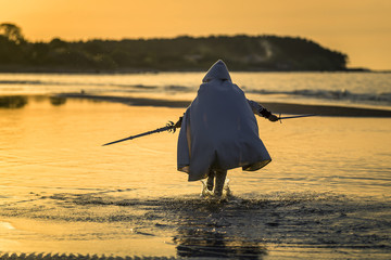 Portrait of assassin in white costume with the sword at the sea. He is posing near water during...