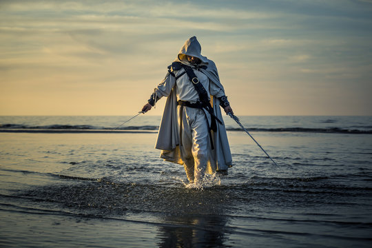 Portrait of assassin in white costume with the sword at the sea. He is posing near water during sunset, soft light.