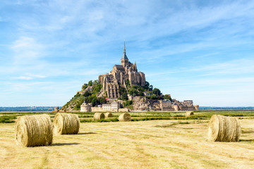 Obraz premium View of the Mont Saint-Michel tidal island, situated in France on the border between Normandy and Brittany, with round bales of straw in a field in the foreground under a blue sky with fibrous clouds.