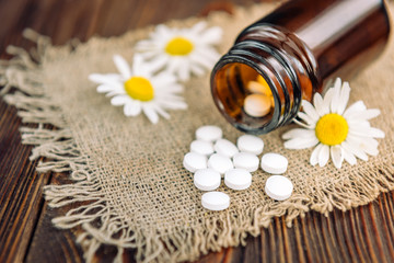 Bottle of white herbal pills and chamomile flowers on dark wooden background, homeopathic medicine.
