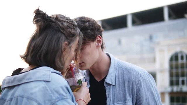 Side view of young couple in blue shirts are dring tea together from a plastic cup using a straw. Woman gently kissing a man. Outdoors