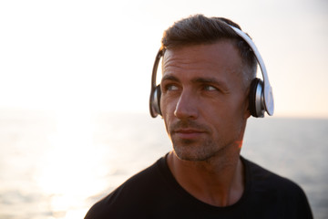 Photo closeup of handsome european man 30s in t-shirt listening to music via wireless headphones, during walk outdoor off coast