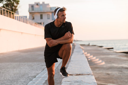 Image of muscular sporty man 30s in shorts and t-shirt walking near seaside, and listening to music via wireless headphones during sunrise