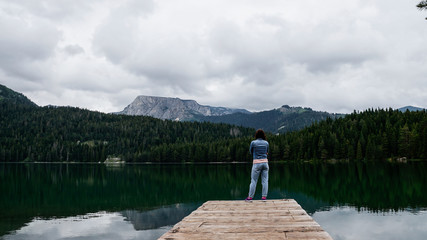 Fototapeta na wymiar The girl stands with her arms crossed in front of her on the pier of black lake in the national Park Durmitor. Montenegro