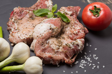 two pieces of steak with spices on black slate, three heads of garlic and tomato, raw meat and green basil leaves, large sea salt