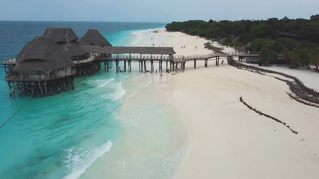 Flight above traditional wooden bungalow with thatched roof and pier bridge on stilts. Wide beach with white sand and azure ocean on cloudy day.  Nungwi and Kendwa beach coastline, Zanzibar, Tanzania.