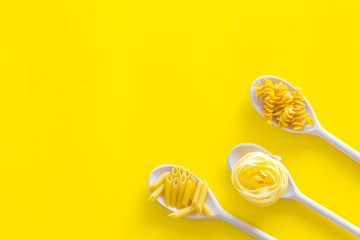 Different raw pasta. Fusilli, penne, fettuccine in spoons on yellow background top view copy space