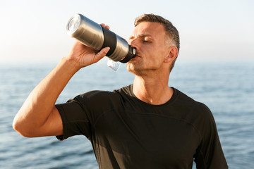 Handsome strong mature sportsman drinking on the beach.