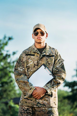 Fototapeta premium portrait of african american soldier in military uniform, cap and sunglasses with notepad in hands