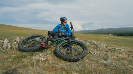 Fat bike also called fatbike or fat-tire bike in summer driving through the hills. The guy sits on a stony sandy and grassy hill after a trip. He rests and enjoys the fresh air.