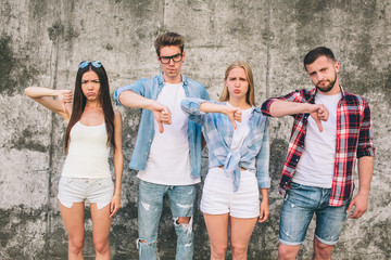 Obraz premium Another picture of unhappy young people standing on grey background. Some of them look a bit angry. They are showing thumbs down. It is dislike symbol.
