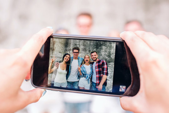 A picture of human's hands holding phone. This human is taking picture of four happy people. They are standing on grey background, posing and smiling.