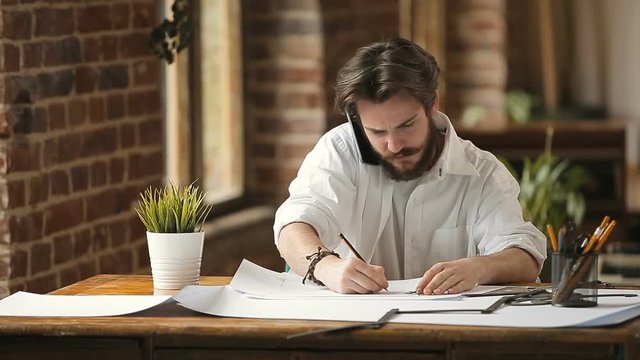 Handsome bearded busy designer working at table workplace, drawings on papers while talking via phone in loft studio