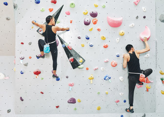 Back view of sportive people in black outfits ascending artificial boulders on clambering wall in...