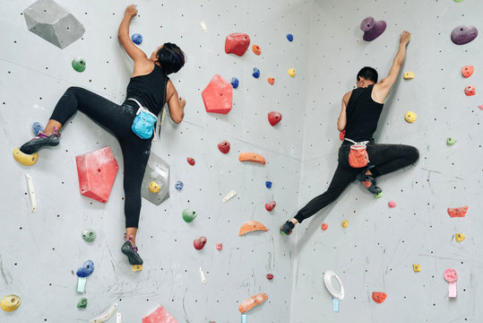 From below shot of athletic man and woman with bags of talk climbing up artificial boulders on wall in gym