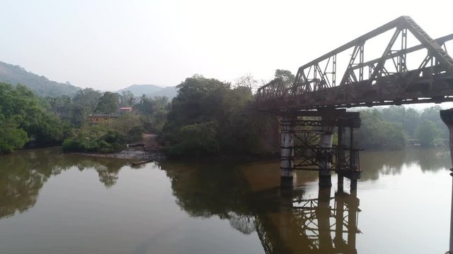 Flying around supports and truss old, rusty, dilapidated bridge over the muddy river on low altitude. Aerial view from a drone.