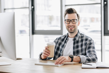 handsome young programmer holding paper cup and smiling at camera while sitting at workplace