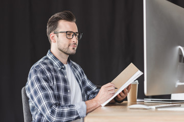 handsome young programmer in eyeglasses taking notes in notebook and using desktop computer