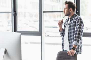 pensive young man holding eyeglasses and looking away while standing at workplace