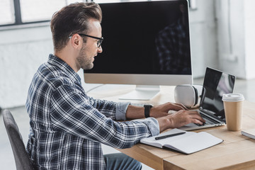 side view of smiling young man in eyeglasses using laptop at workplace